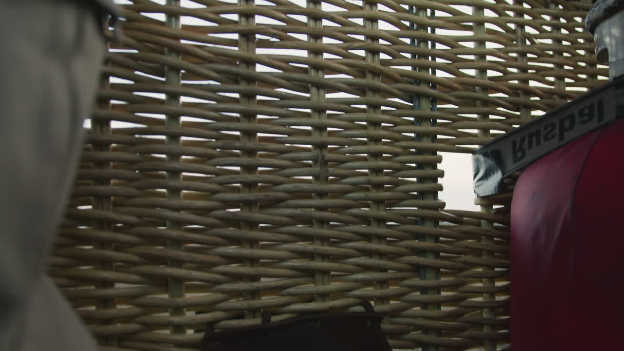 close up of woven basket side from hot air balloon gently floating above open fields during sunset with soft sky tones in background and visible harness strap
