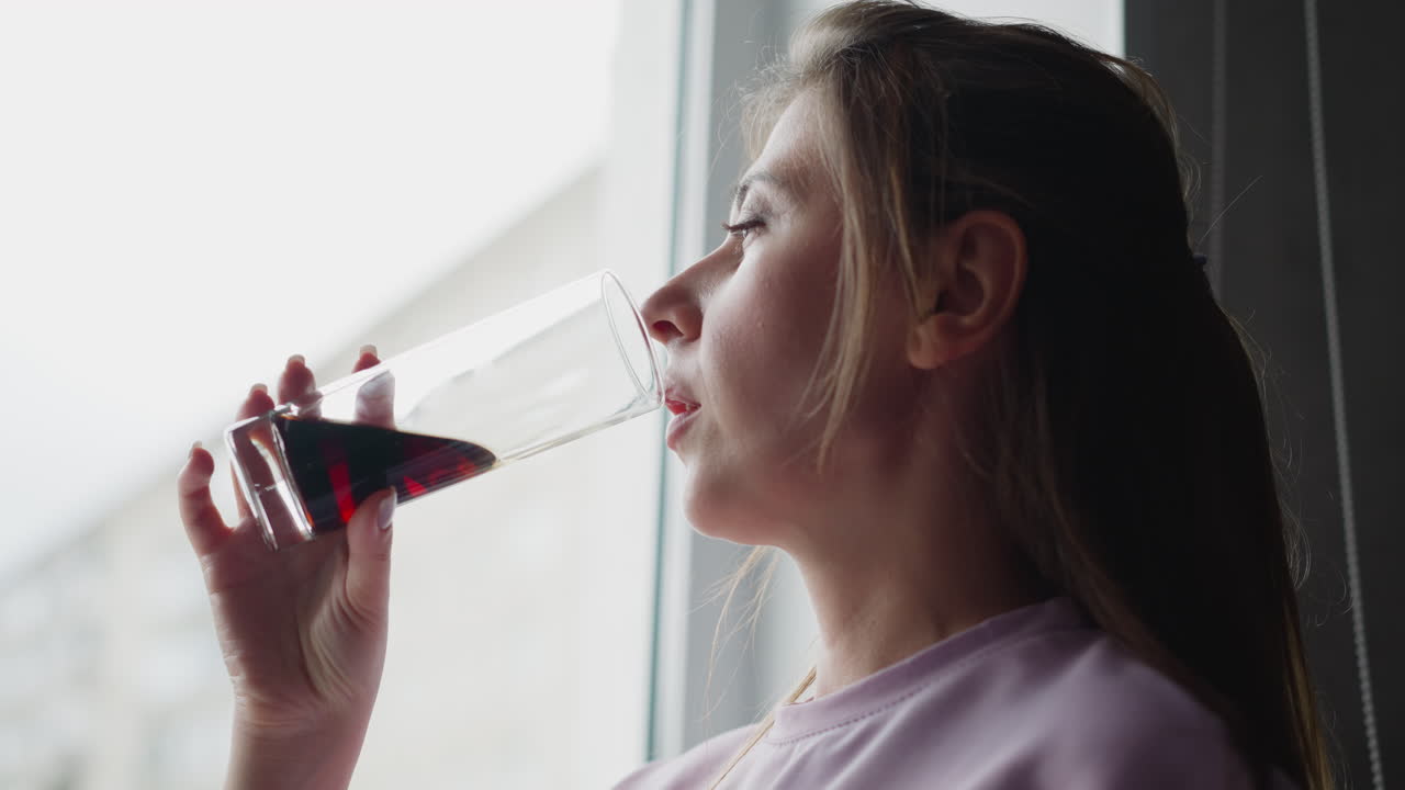 close up elegant young woman drinking wine near window indoors bright natural daylight illuminating face relaxed lifestyle enjoying beverage peaceful morning moment casual home leisure
