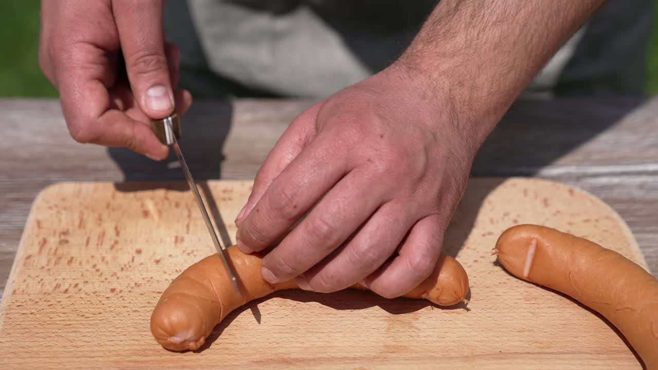 Man's hands prepare sausages for barbeque. Chef in doing incisions on juicy sausages with a knife on a wooden board before grilling the outdoors. Close-up.