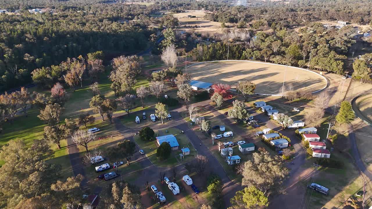 Drone footage glides above a quiet residential neighborhood surrounded by trees and open fields, with long shadows and warm, late afternoon sunlight