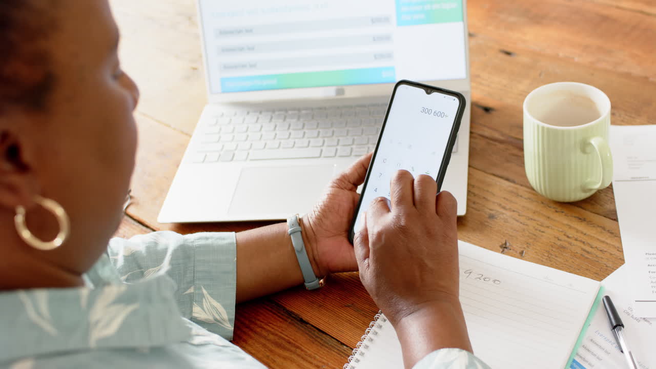 mujer afroamericana de edad avanzada haciendo papeleo, usando portátil, teléfono inteligente en una habitación soleada, cámara lenta