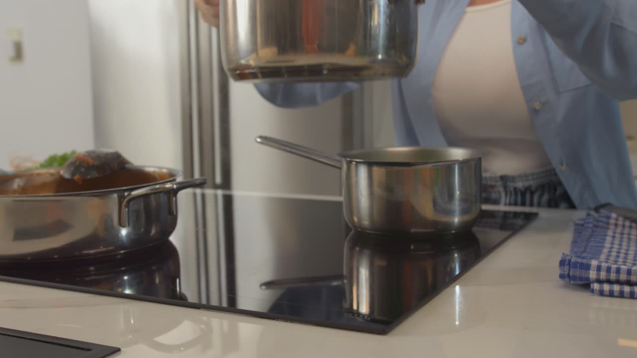 A woman is cooking in a kitchen with a pot on the stove. She is wearing a blue shirt and a white shirt