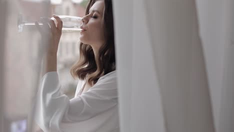 woman in a white robe drinking champagne by a window