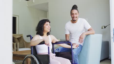 Happy-biracial-woman-in-wheelchair-and-smiling-male-partner-sitting-holding-hands-in-living-room