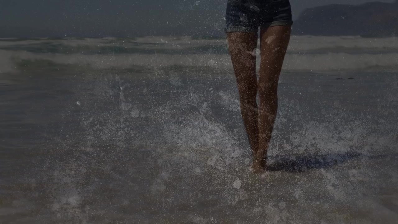 Vrouw loopt door de ondiepe branding op het strand, haar spijkerbroekje spat water op rond haar enkels.