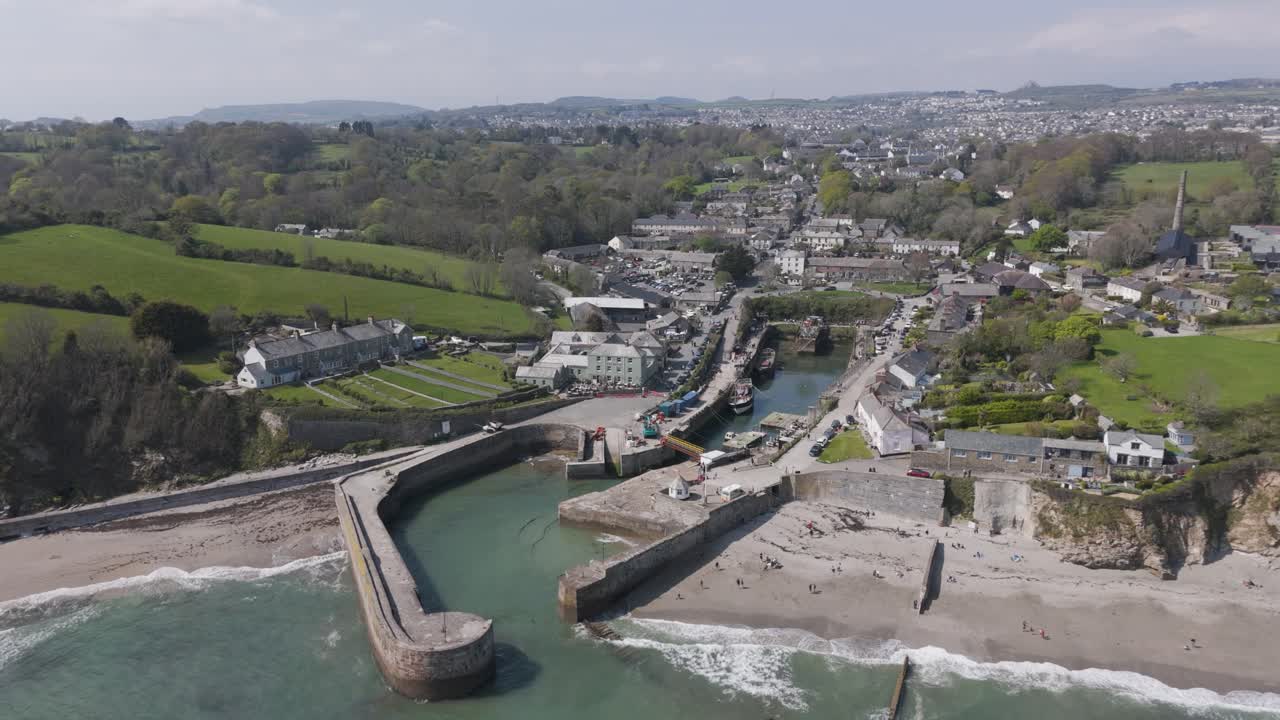 Aerial orbit of Charlestown harbour with stone quay, tall ships and traditional cottages surrounding sandbanks at low tide against rolling green hills