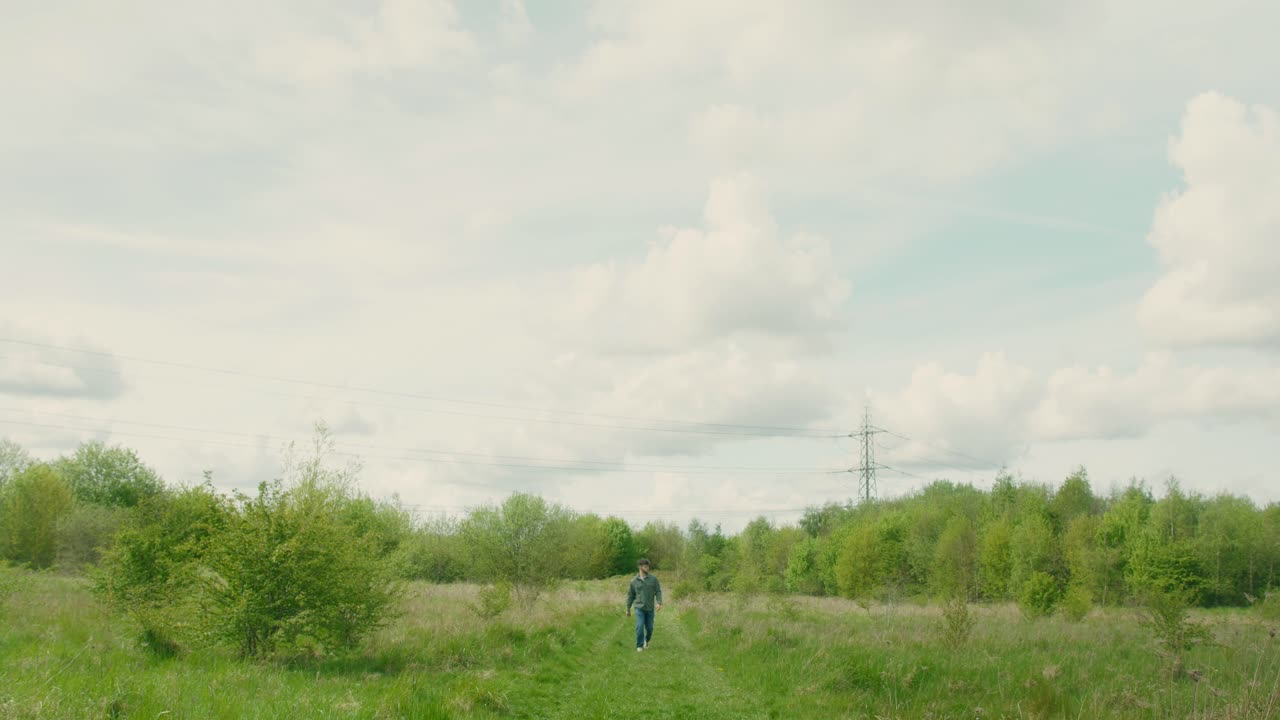 Man walks slowly through tall grass under soft cloudy sky, horizon wide and peaceful, nature surrounds him, wide angle overview