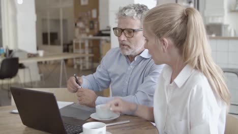 bearded serious ceo in glasses working on laptop, writing notes with her assistant next to him