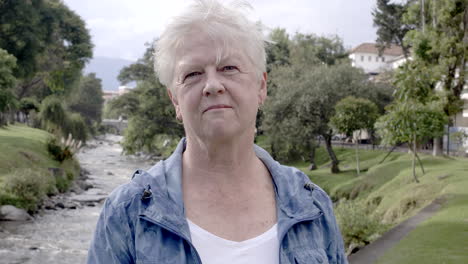 anciana sonriendo a la cámara junto a un río y una calle en la parte de atrás