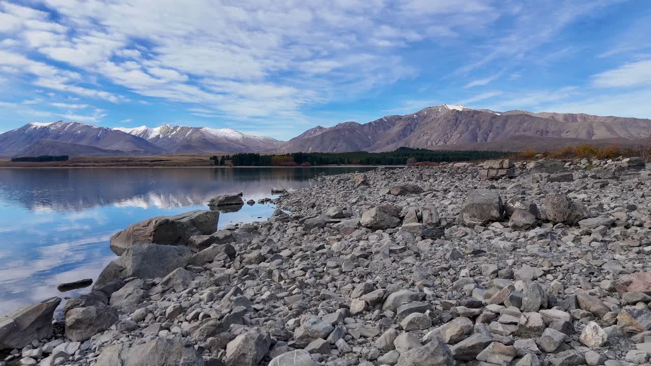 Aerial footage captures Lake Tekapo's rocky shoreline with clear reflections and mountainous backdrop under a bright blue sky