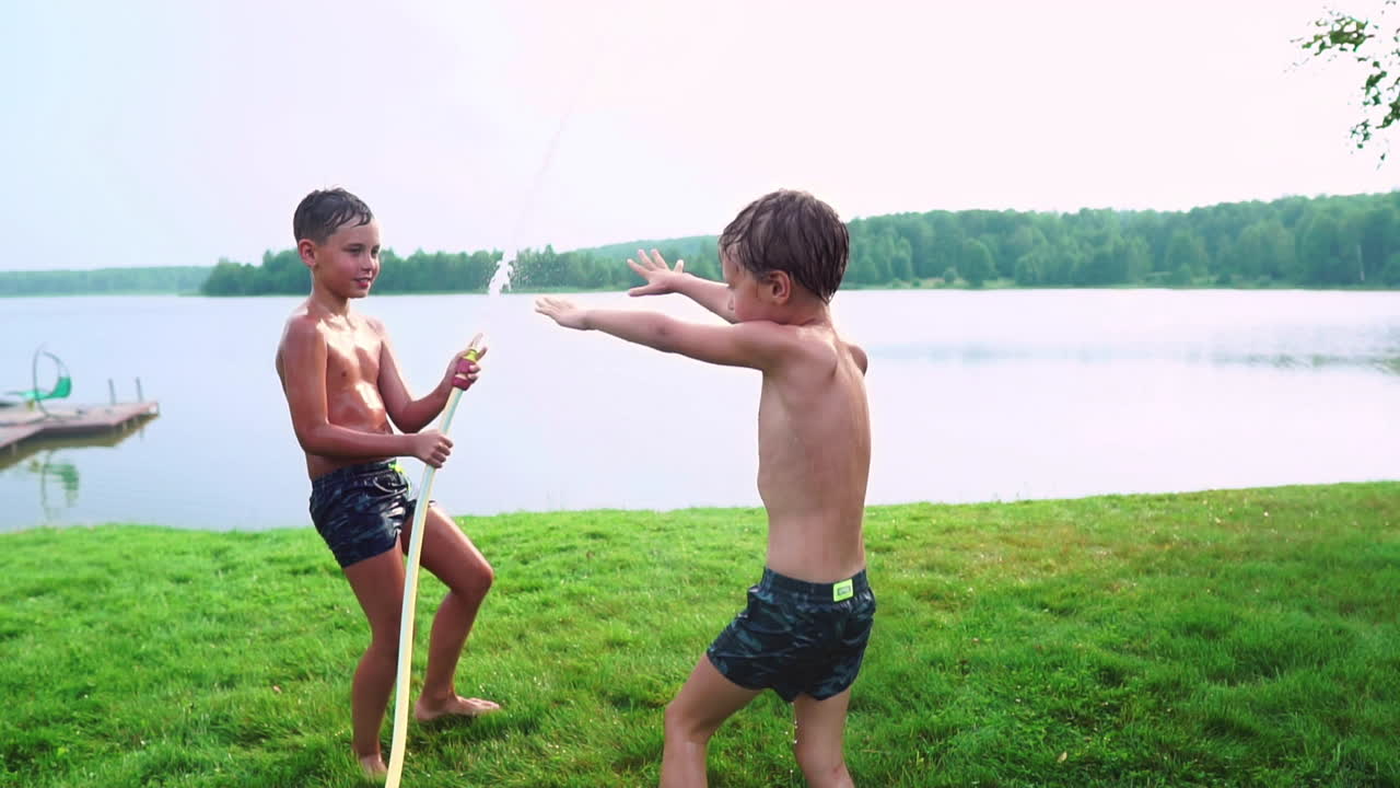 여름에 수영복을 입은 소년은 동생에게 물을 부어 호수 근처의 잔디 ⁇ 에서 공원에서 재미를 느끼고 있습니다.