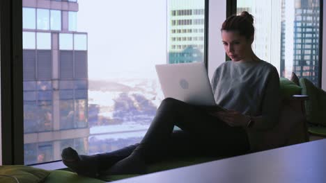 una mujer atractiva está sentada junto a la ventana con vista al centro de negocios, abriendo su portátil y comenzando a escribir en un teclado