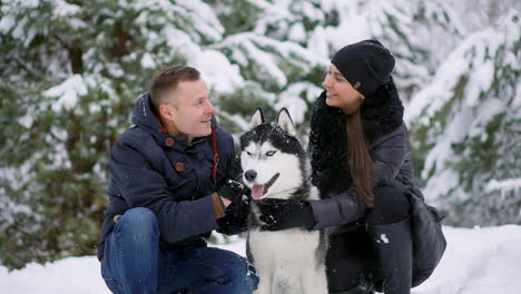 a man and a woman sitting hugging a dog siberian husky in the winter forest smiling and looking at each other and at the camera. slow motion happy family