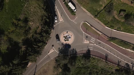 A-traffic-roundabout-surrounded-by-greenery-on-a-sunny-day,-featuring-moving-vehicles-and-distinct-road-markings,-aerial-view