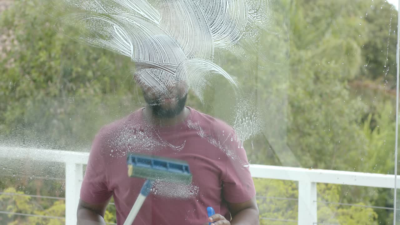 Bebaarde man spuit met blauwe fles en trekt balkon glasplaat schoon om schuim te verwijderen.