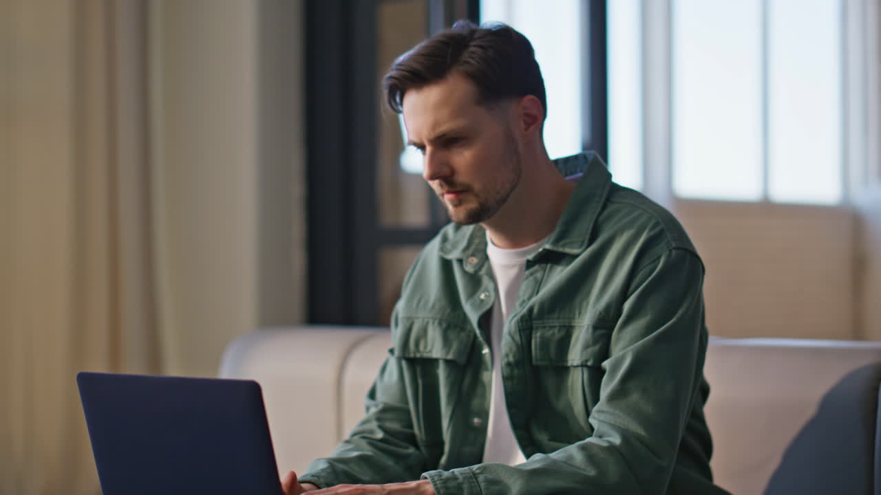 Focused man working laptop at windows apartment closeup. Pensive online student