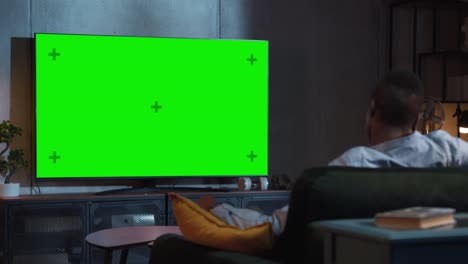 black man watches green mock-up screen tv while sitting on a couch at home. male stream evening movie. over the shoulder view. entertainment concept