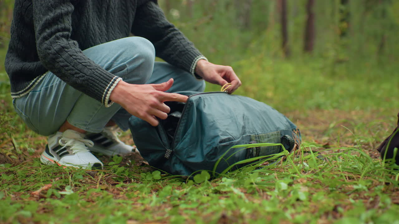 camper squatting on forest ground unzipping green outdoor bag and removing camping equipment with partial view of second bag nearby surrounded by greenery