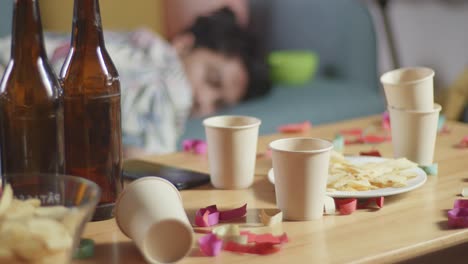 close up of four empty bottles of alcohol with paper cups, snack, and smartphone are on the table while a man sleeping on a sofa for celebrating party at home