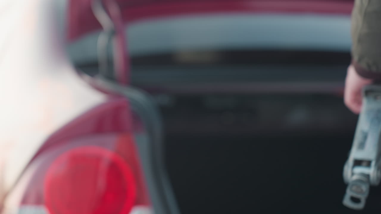 Close up of person in winter jacket holding metal jack and spanner beside open car boot, preparing to close trunk on snowy day, red car taillight and soft daylight reflections visible on body