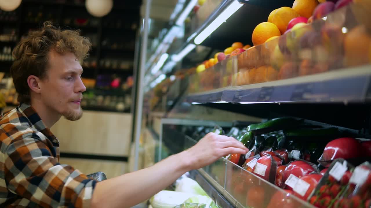 un hombre caucásico con camisa a cuadros comprando frutas y verduras en el departamento de productos de un supermercado de una tienda de comestibles. eligiendo tomates frescos perfectos del estante inferior. vista lateral