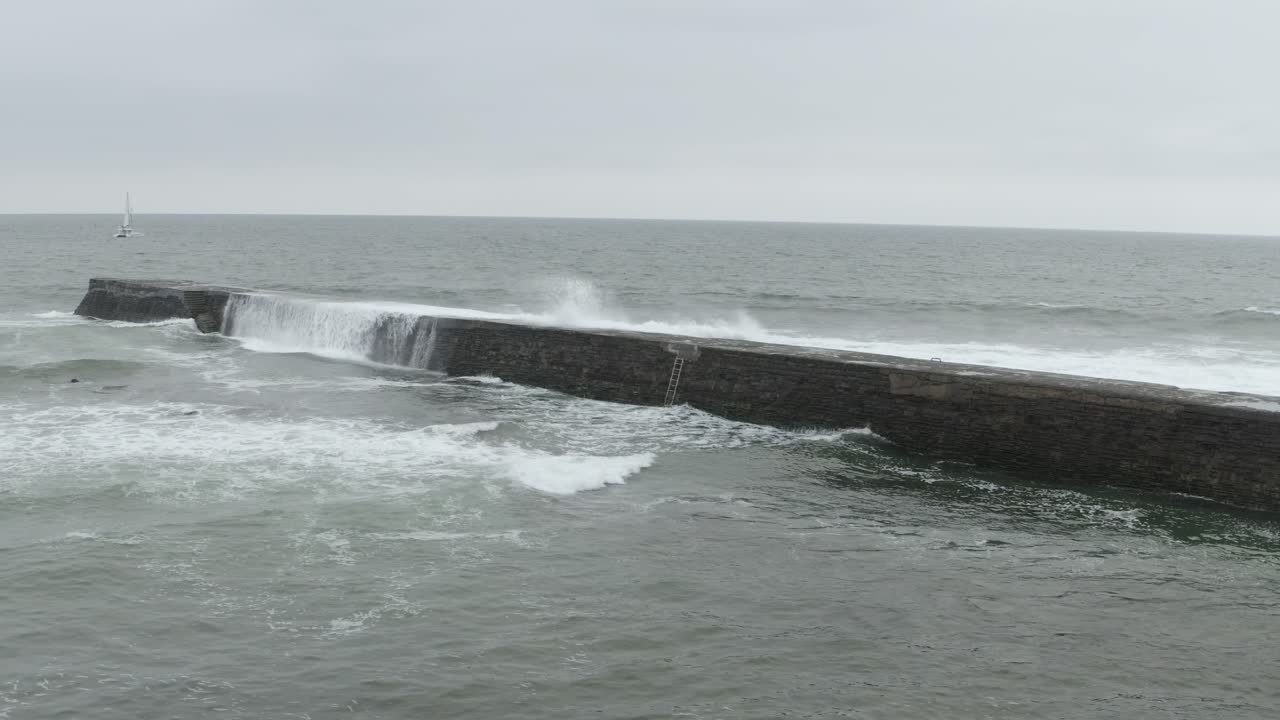 Waves crashing over seawall, sailboat in distance, Saint-Jean-de-Luz, France. Aerial drone, low flight