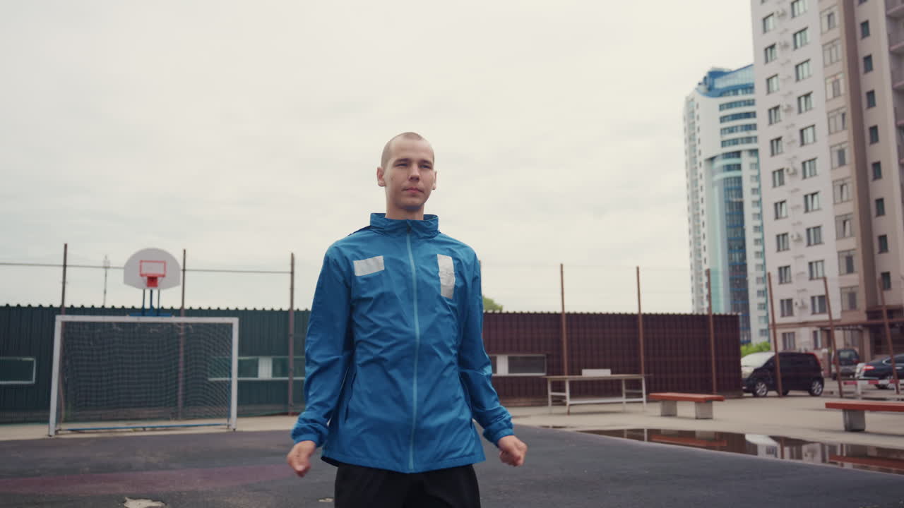 city playground white male warming up in blue jacket, soccer ball at feet and goal in background, fists pump in subtle celebration, skyscraper and tower silhouettes, focused pregame routine