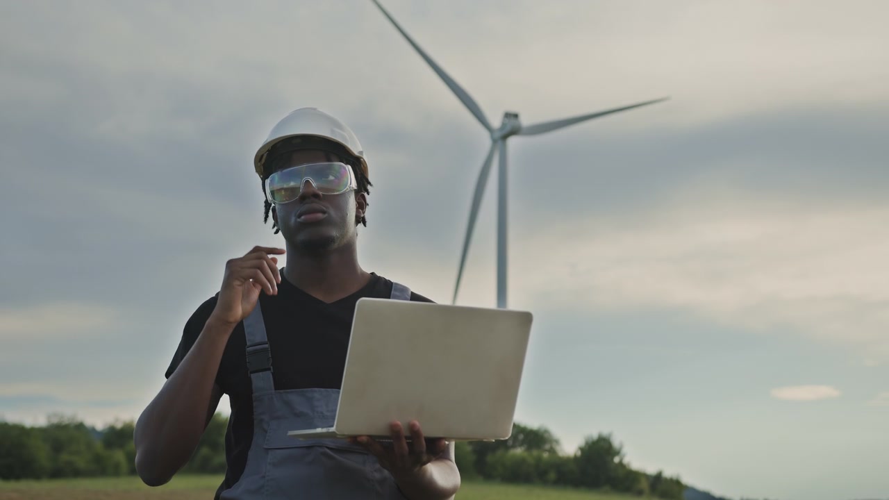 Ingegnere afroamericano con cappello e occhiali di sicurezza controlla i dati sul laptop vicino a una turbina eolica