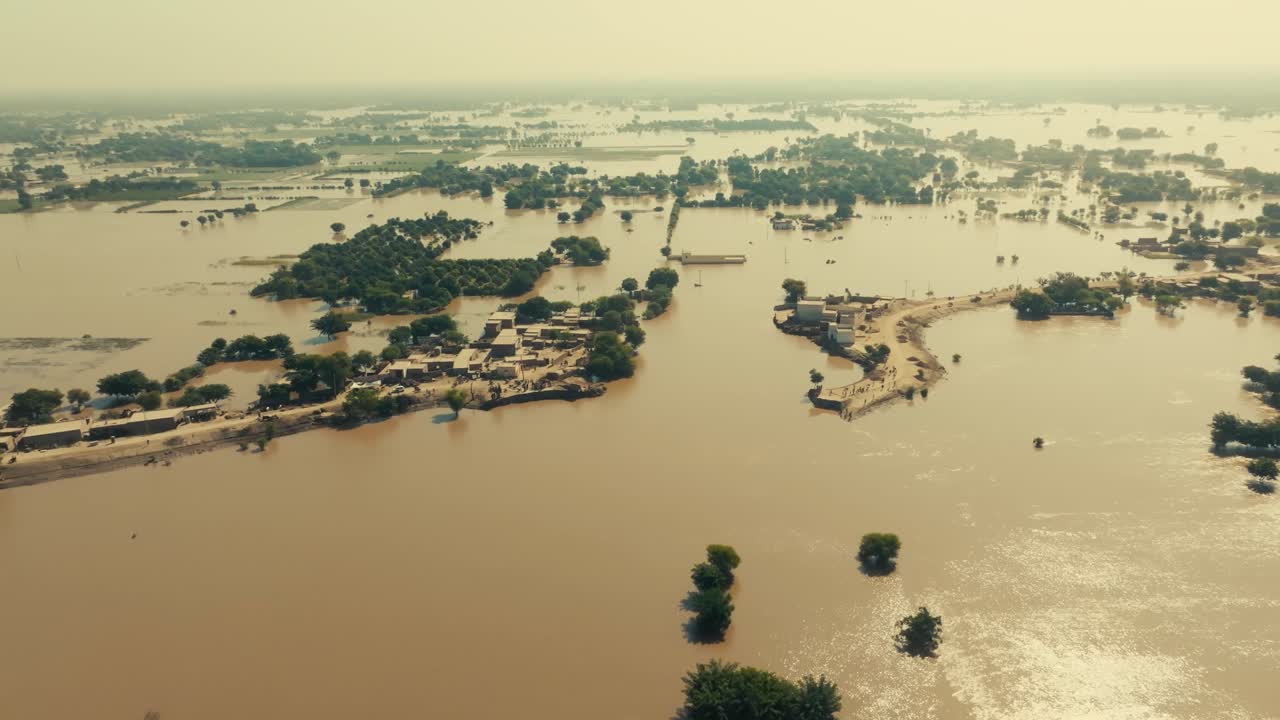 High aerial orbit shot over flood-damaged fields and a small, illuminated residential area on a piece of dry land in Jalalpur Pirwala, under a sepia-like sunny sky