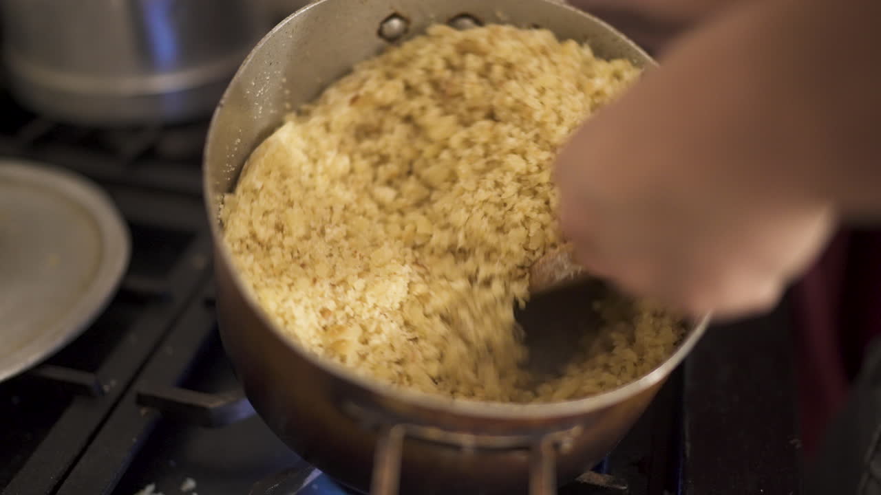Reviro is stirred with a wooden spoon while cooking on a stove, showing golden crumbs forming in the metal pan — traditional rural South American preparation captured in kitchen close-up.