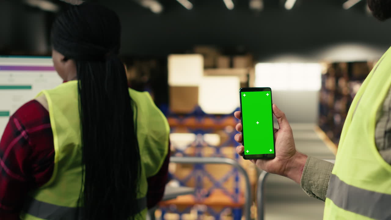 Warehouse Worker Holding Green Screen Smartphone While Colleague Works on Computer