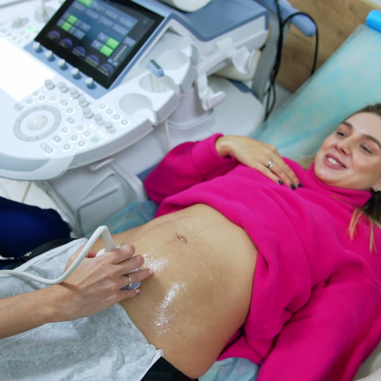 Smiling Caucasian woman in pink sweater is checked on the ultrasonic machine. Woman is happy to see her future child on the monitor. Top view