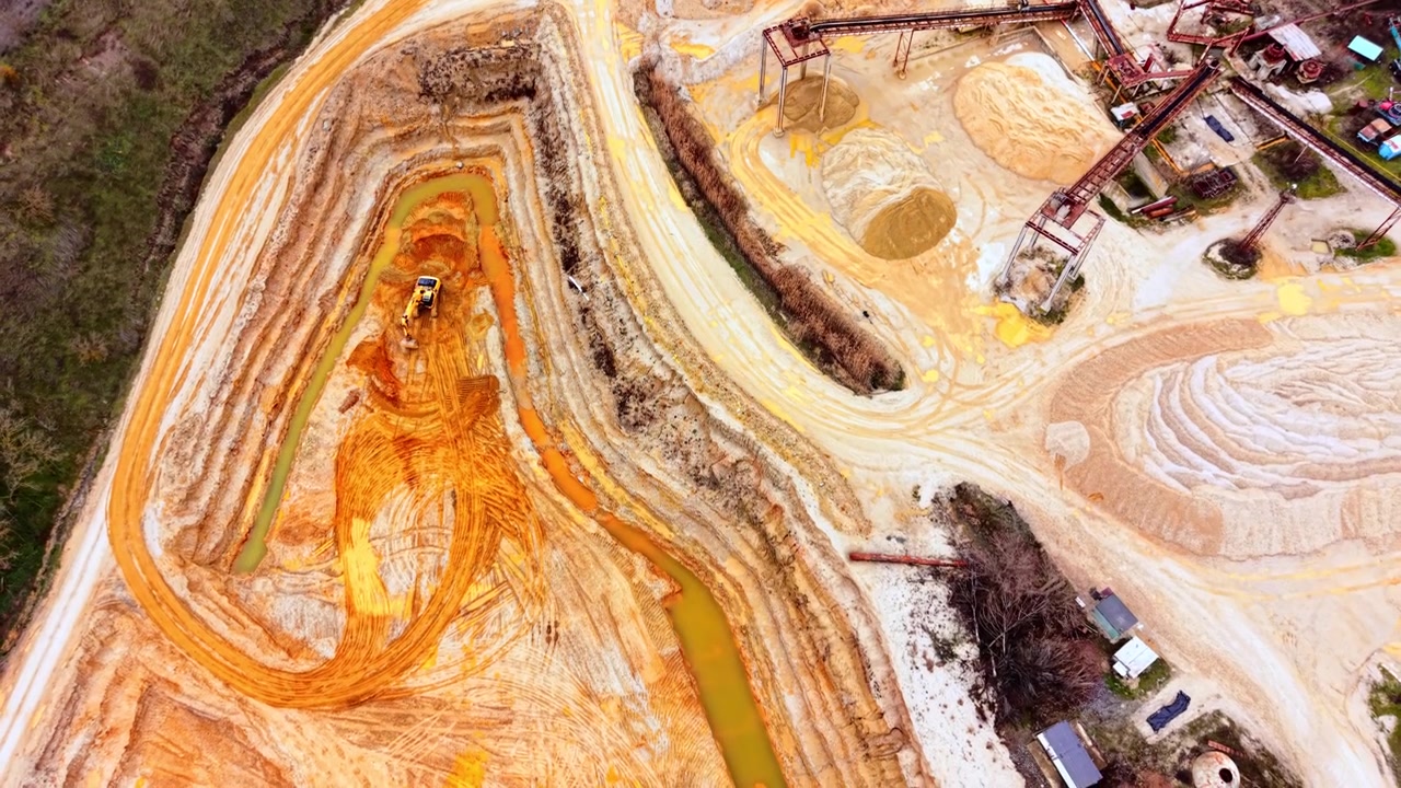 Sand quarry pit with excavator near forest. Aerial view of terraced orange sand banks, a winding stream, and processing equipment.
