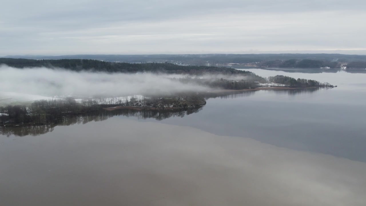 Luftfoto med drone, der viser søen Anten nær Gräfsnäs med tåge svævende over kystlinjen og roligt vand omgivet af vinterlandskab i Alingsås, Sverige.