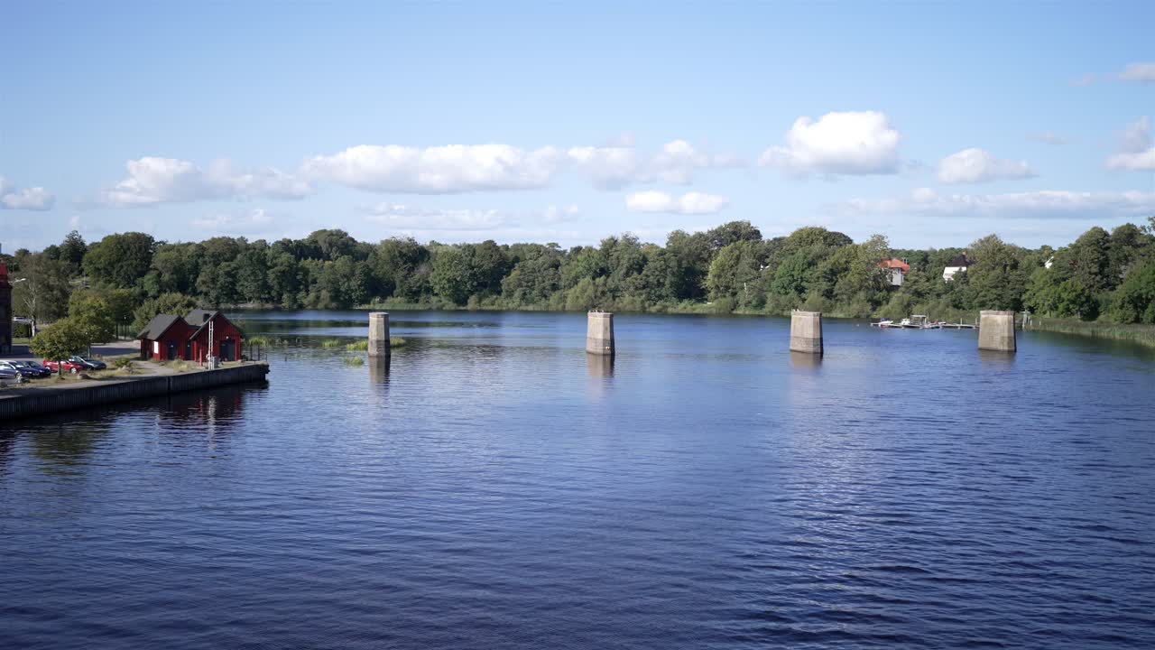 A quiet Swedish lake with large concrete bridge supports standing in the water, but no bridge above. A red cabin sits on a dock nearby. A peaceful, atmospheric scene.