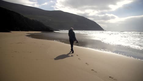 Fotógrafa-Camina-En-Una-Playa-De-Arena-En-Irlanda,-Dejando-Huellas-Atrás,-Olas-Golpeando-La-Orilla,-Cielo-Soleado-Con-Nubes,-Toma-En-Cámara-Lenta