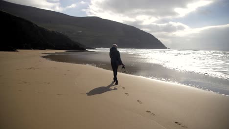 fotógrafa camina en una playa de arena en irlanda, dejando huellas atrás, olas golpeando la orilla, cielo soleado con nubes, toma en cámara lenta