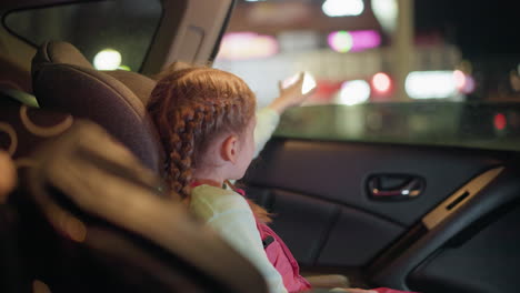una vista cercana de una niña jugando con su mano a través de la ventana del coche, con una vista borrosa de las luces al otro lado de la carretera, la niña, con el cabello trenzado, está sentada en el coche