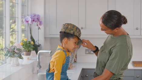 madre del ejército estadounidense en uniforme en casa de licencia abrazando a su hija con su gorra en la cocina de la familia