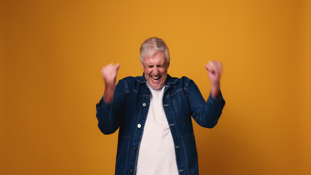 A senior man in a denim jacket posing against a yellow background