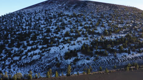 Aerial:-Snow-covered-mountain-with-pine-trees-at-Sunset-Crater,-Flagstaff---drone-backward-shot