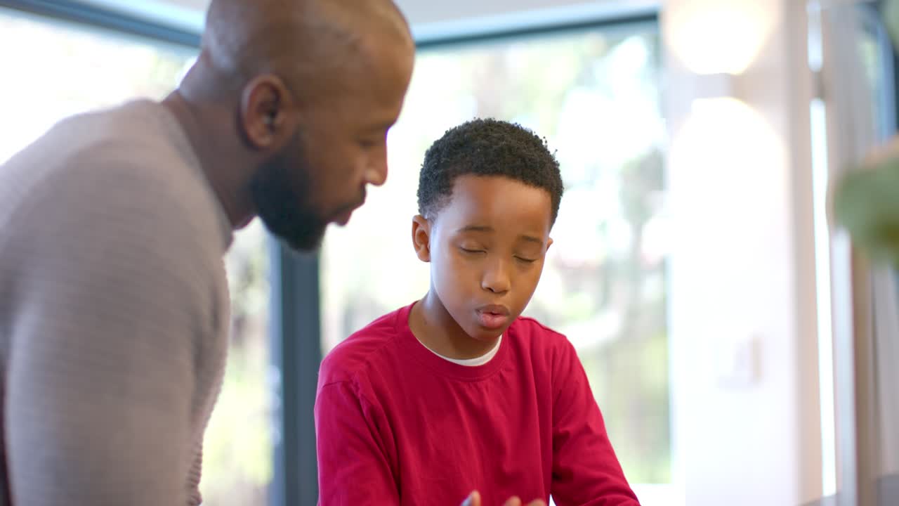 African American father guiding son adjusting pencil, writing homework in notebook at dining table
