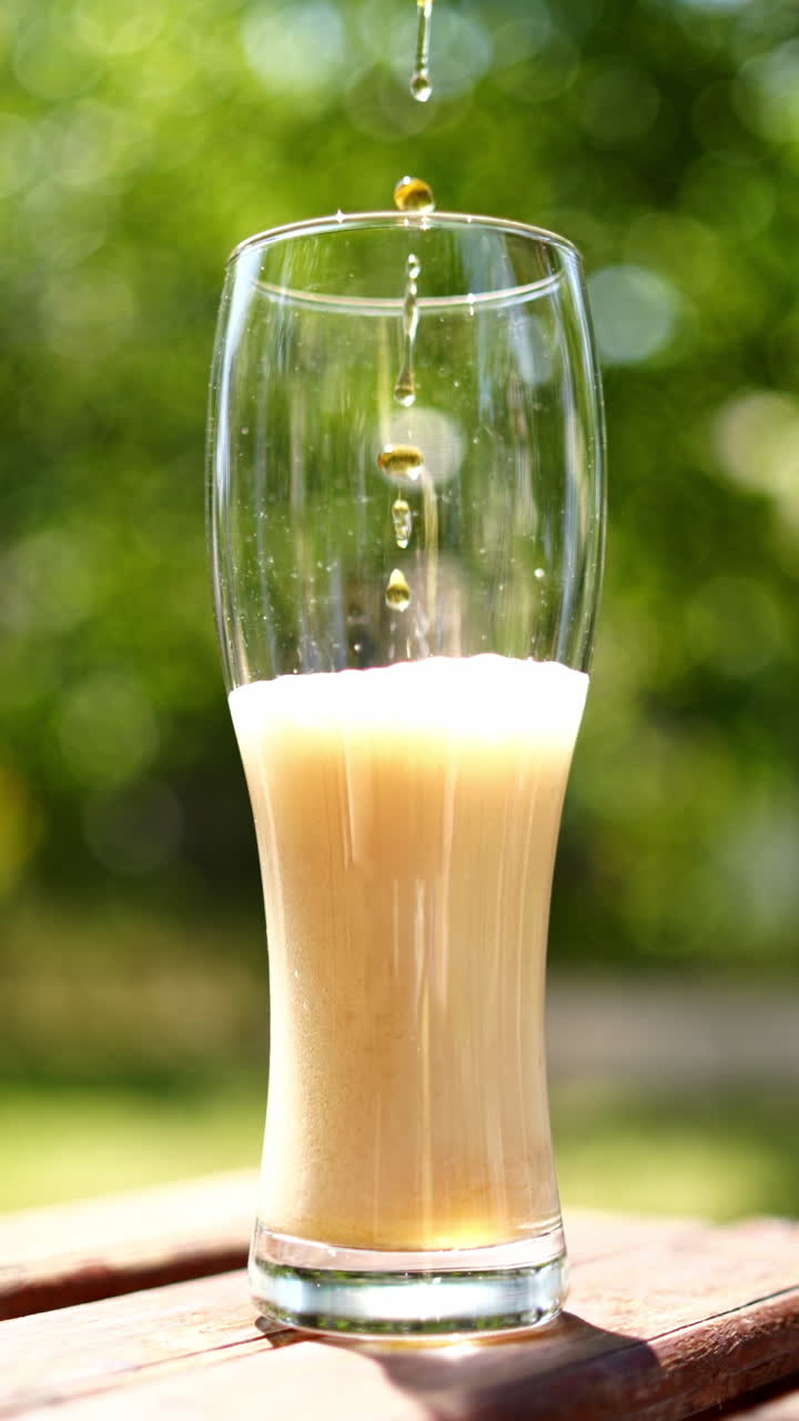 Process of pouring beer to a tall glass on a wooden table. Beer is pouring from the top into the glass with forming wavesfrom the foam. Close-up. Vertical video