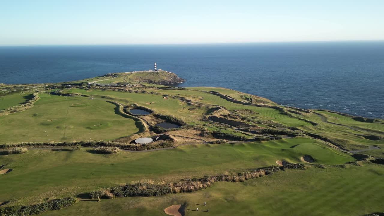 Green headland pointed to calm blue Atlantic Ocean, with protruding lighthouse. Old Head of Kinsale golf course, county Cork, Ireland aerial.