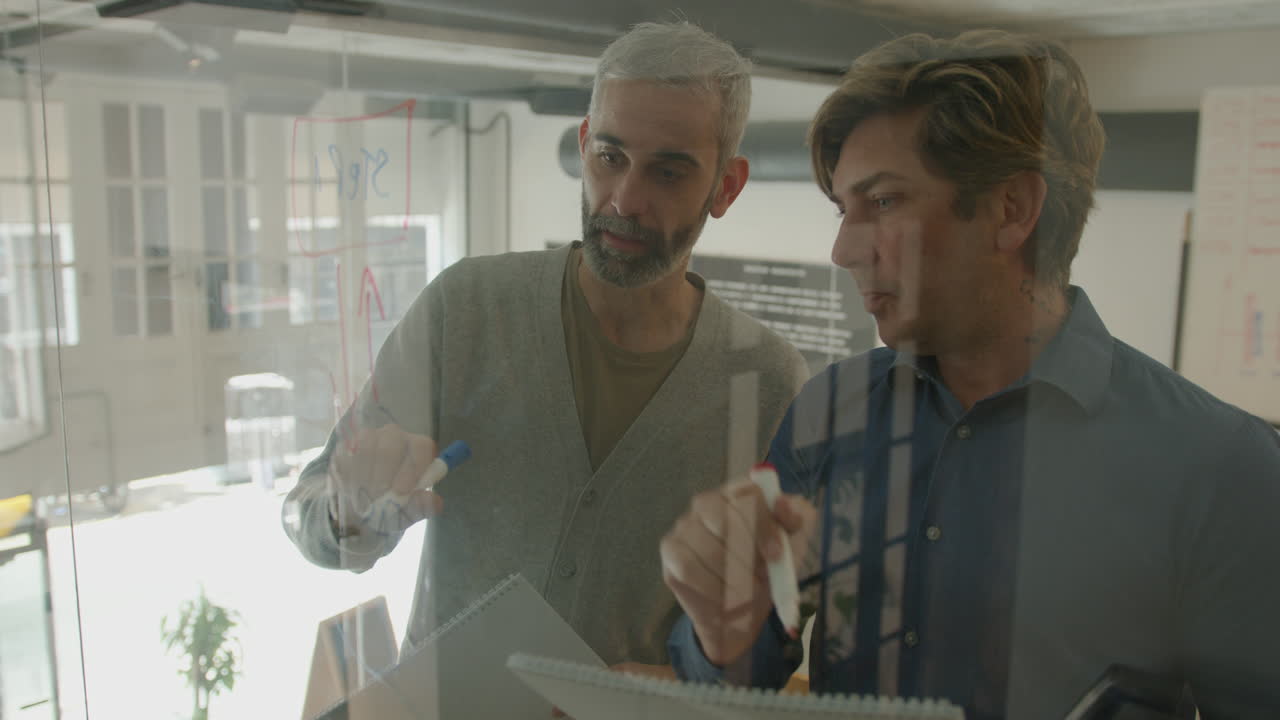 Senior Businessman Discussing Scheme on Glass Office Wall with Colleague