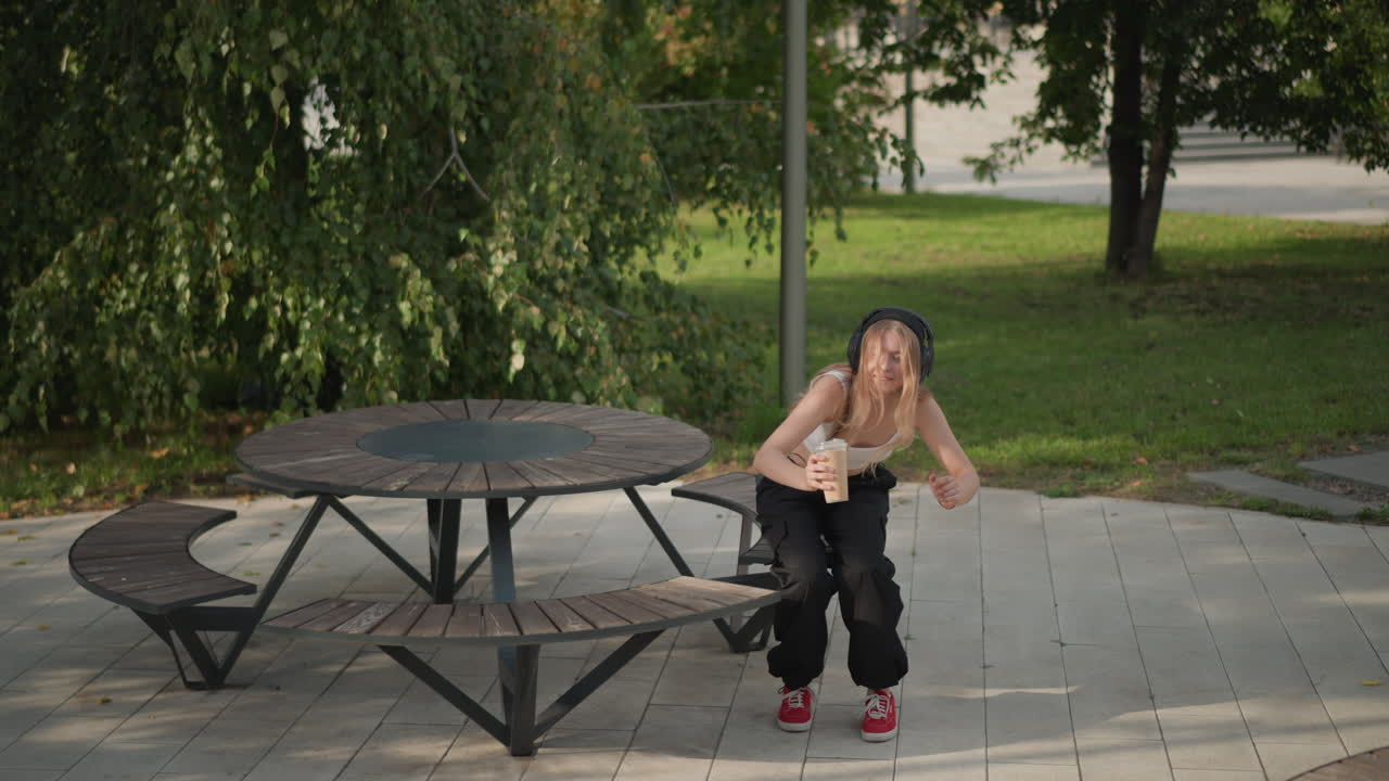 young woman street park sipping coffee while dancing near picnic table, playful leg resting on bench, red sneakers and loose black pants, tank top, shaded afternoon, relaxed rehearsal vibe, candid