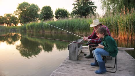 side view of a teen boy sitting with his grandfather on the lake pier, talking and holding a fish on the fishing rods while fishing together