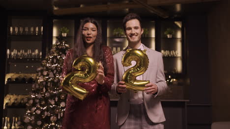 donna e uomo che indossano abiti eleganti tenendo palloncini con i numeri 22 mentre ballano alla festa di capodanno