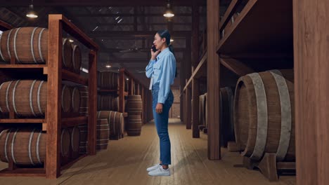 full body side view of an asian female professional worker standing in cellar of winery, she is speaking with someone in the phone