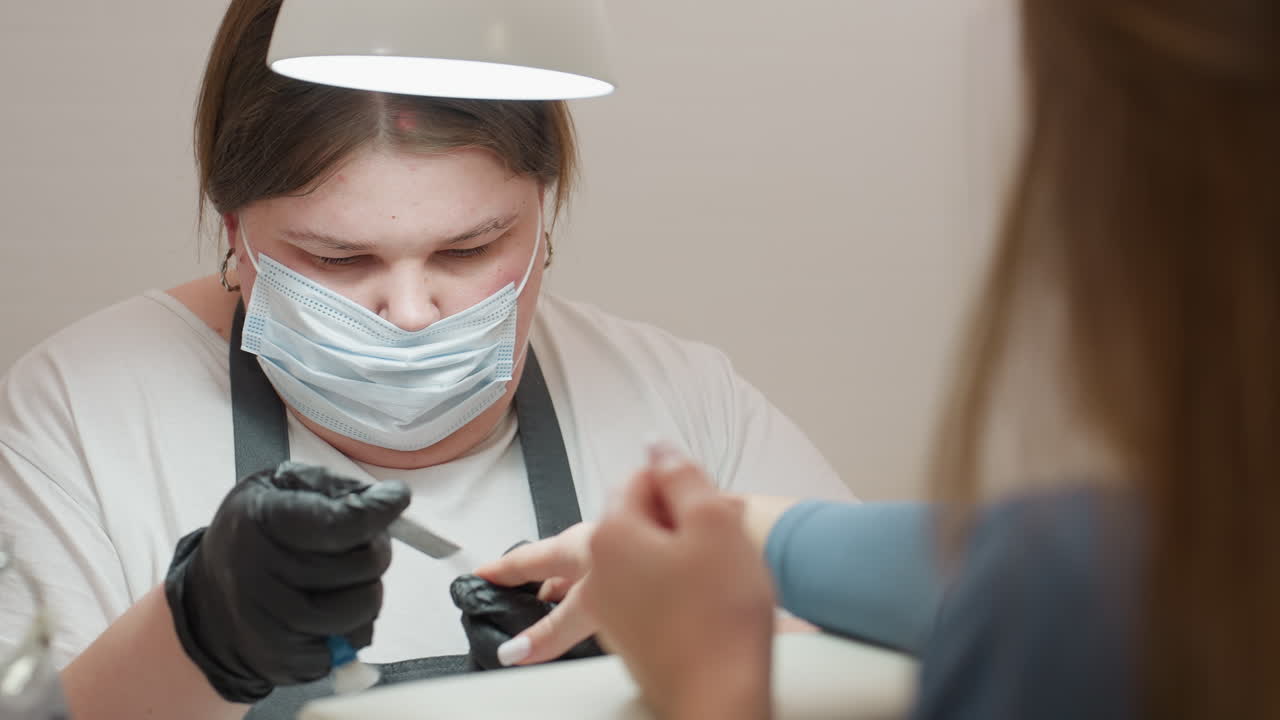 Nail technician in white polo and apron carefully files client nails while using blue brush for cleaning and client observes process attentively, highlighting personal care and salon hygiene